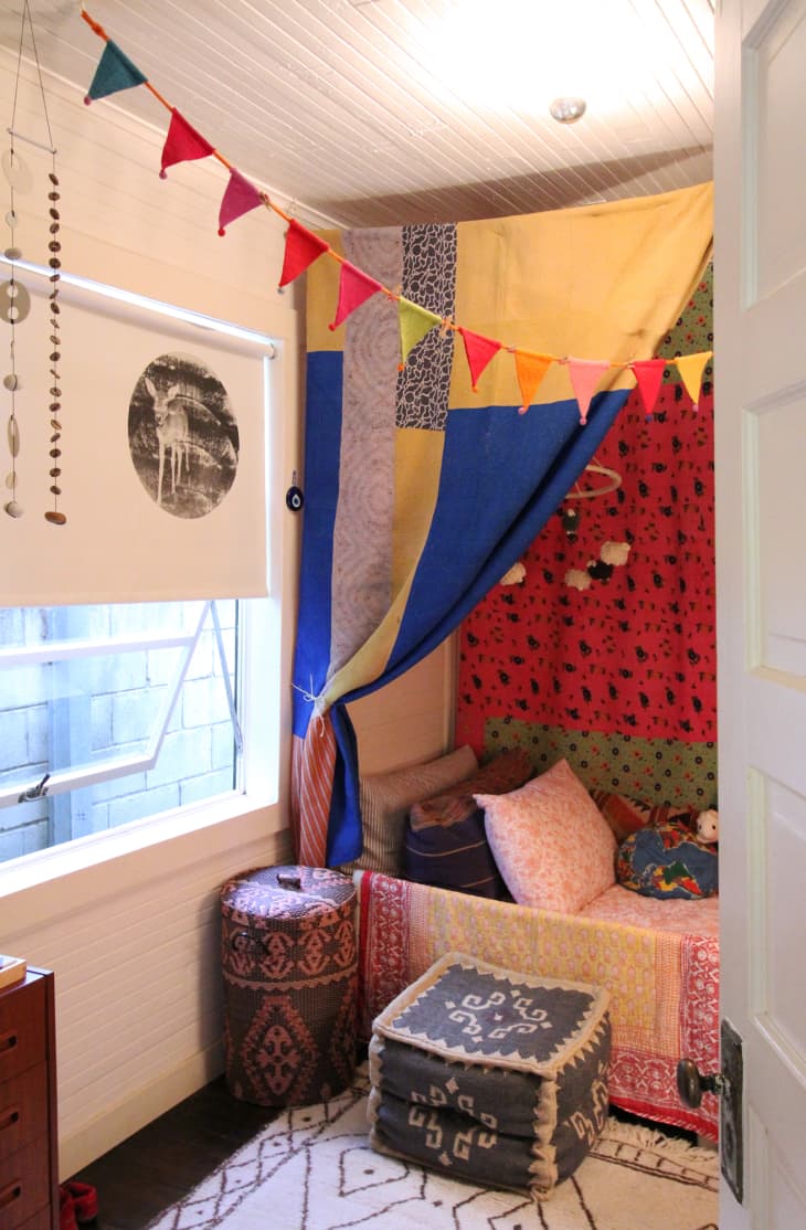 Cozy nook with colorful patchwork canopy, patterned cushions, and bunting flags, next to a window with a decorative blind.