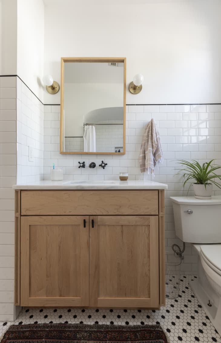 Bathroom with wooden vanity, white subway tiles, round mirror, brass sconces, and a potted plant on the toilet.