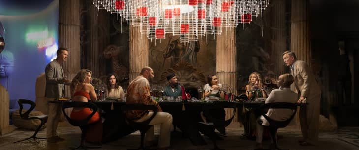 A lavish dining scene with eight people seated around a large black table, ornate chandelier above, and colorful decor.