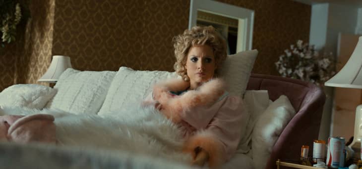 Woman in pink robe with fur trim sitting on a plush bed, surrounded by pillows and lamps, in a vintage-style room.