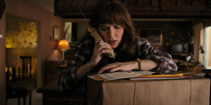 Woman in plaid shirt talking on a cordless phone, leaning on a large cardboard box in a dimly lit room.
