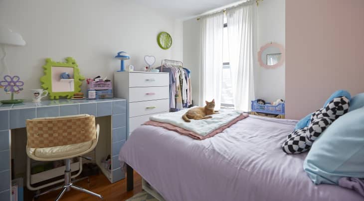 A bedroom with one pink feature wall. A blue tiled desk is next to a white dresser. On the bed is a purple duvet with an orange cat laying down on it.