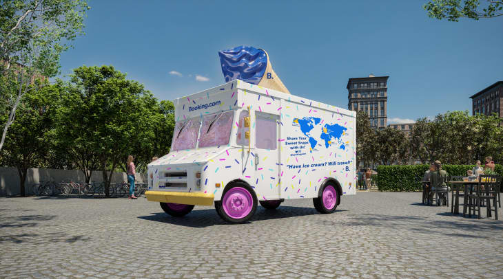 Ice cream truck with sprinkles and a large cone on top, parked in a sunny plaza with trees and people nearby.