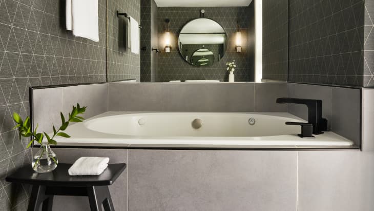 Modern bathroom with a large bathtub, black faucet, geometric tile walls, and a small table with a plant and towel.