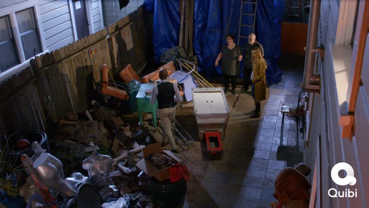 Cluttered backyard with four people discussing amidst scattered furniture, mannequins, and a blue tarp.