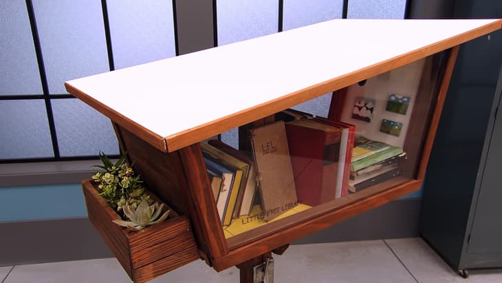 Wooden little free library with books, a white top, and a side planter with succulents.