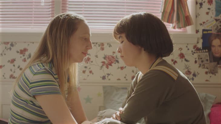 Two people sitting closely in a floral wallpapered room with pink blinds and posters on the wall.