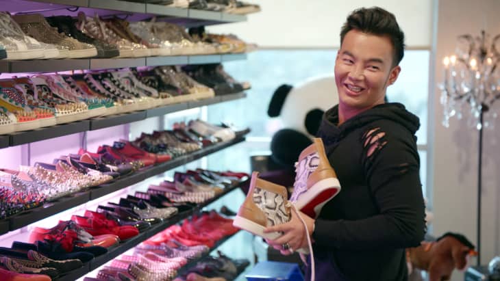 Man holding sneakers in a room with shelves of colorful designer shoes and a large panda plush in the background.