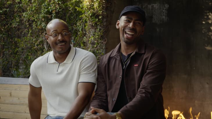 Two men sitting outdoors, one in a white shirt and glasses, the other in a dark jacket and cap, smiling near a fire pit.
