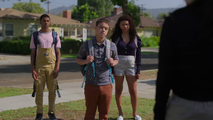 Three teenagers with backpacks standing on a suburban street, facing an unseen person.