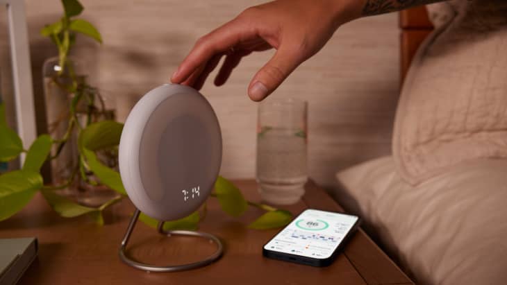 Hand reaching for a round digital clock on a bedside table with a smartphone, glass of water, and green plant.