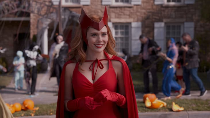 A woman in a red costume with a cape and headpiece smiles, surrounded by people in Halloween costumes and pumpkins.