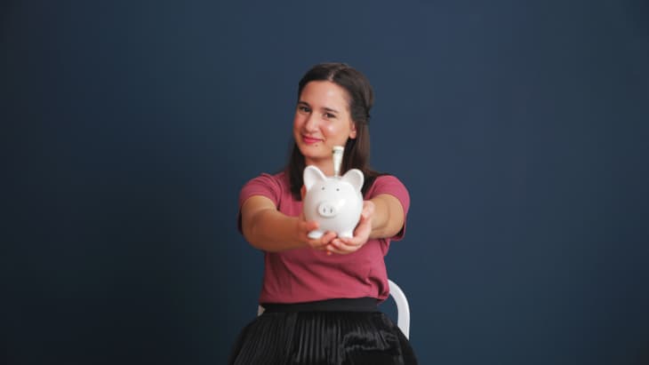 Woman in a pink shirt holding a white piggy bank against a dark blue background.