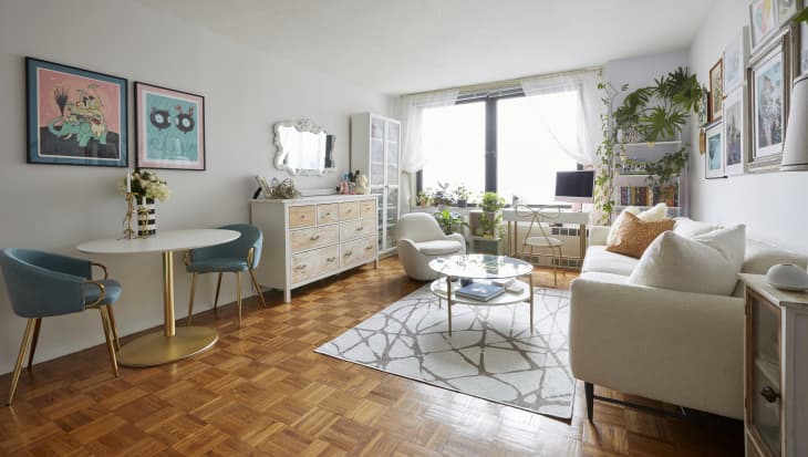 Bright living room with white sofa, round glass coffee table, plants, and colorful wall art.
