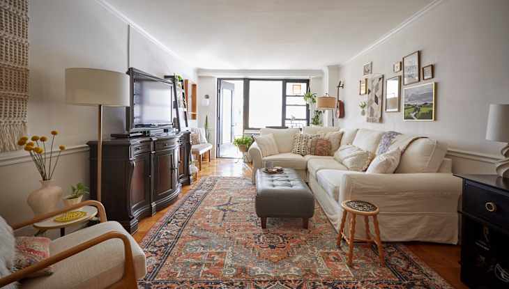 Living room with white sectional sofa, patterned rug, dark wood TV stand, and wall art, leading to a bright windowed area.