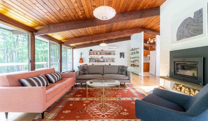 wood slated ceiling, wood beams, angled ceiling, red pattern rug, pink couch, blue arm chair, white brick fireplace, black metal inlaid fireplace, guitar, floating bookshelves, round glass coffee table