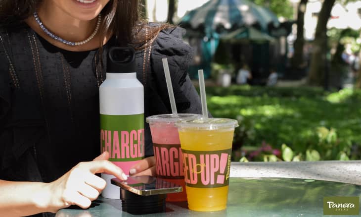 Woman in black dress using smartphone at outdoor table with two colorful drinks and a water bottle labeled "Charged Up!"