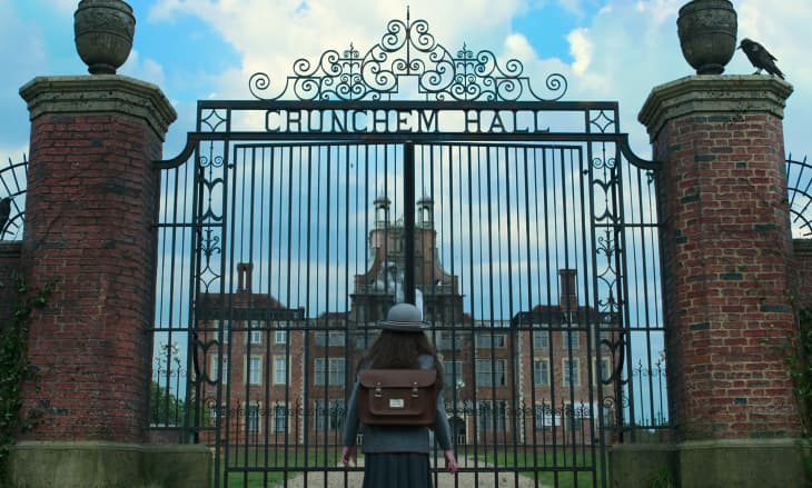 A person with a brown backpack stands before ornate iron gates labeled "Crunchem Hall," with a historic building visible behind.