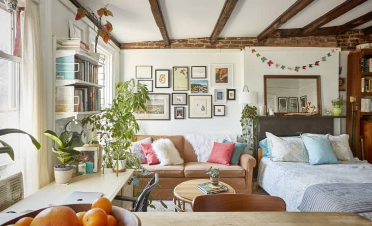Cozy living room with a tan sofa, colorful pillows, plants, bookshelves, and a gallery wall under exposed wooden beams.