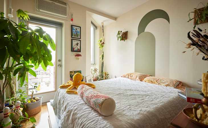 Stuffed octopus on bed against decoratively painted wall in bedroom area with framed New Yorker art and potted plants.