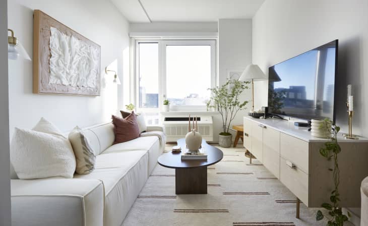 A narrow living room with a white couch, light wood TV stand, and a large window