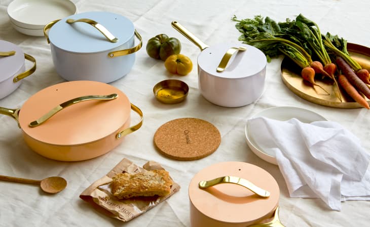 Colorful cookware with gold handles, fresh carrots, tomatoes, and bread on a white tablecloth.