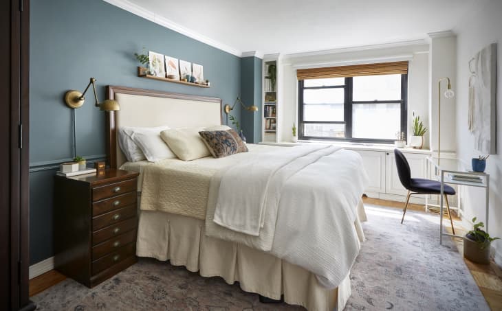 Cozy bedroom with teal accent wall, white bedding, wooden nightstand, and a desk by the window with plants and decor.