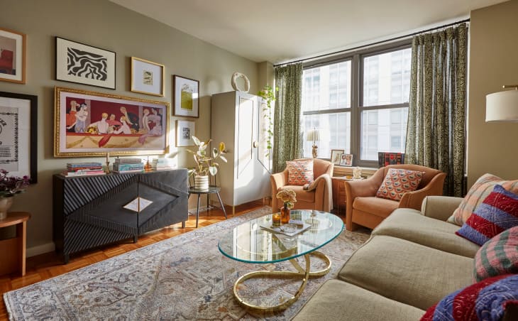 Living room with sage/olive colored walls, gallery wall, black credenza, 2 peach armchairs with throw pillows, gray patterned rug, gray wardrobe/cabinet, patterned floor to ceiling curtains, sofa with patterned throw pillows, glass and gold oval coffee table