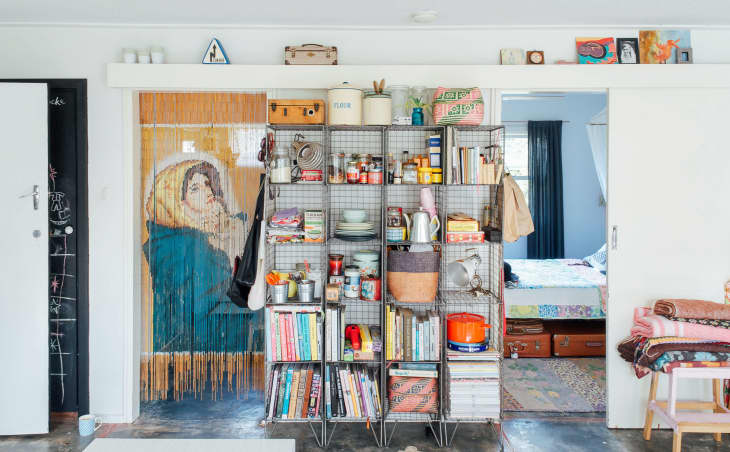 Open shelving with books, kitchen items, and decor in a room with a beaded curtain and view into a bedroom.