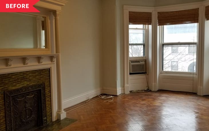 Empty living room with a decorative fireplace, parquet floor, and three large windows with bamboo blinds.