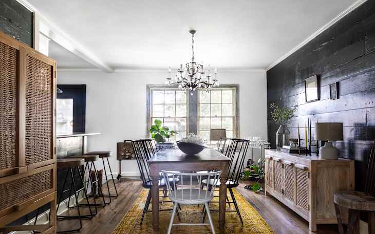 Dining room with wooden table, mixed chairs, chandelier, black accent wall, sideboard, and plants on a yellow rug.