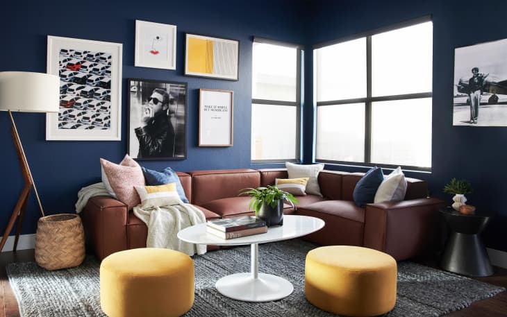 Navy blue living room with brown sectional sofa, yellow ottomans, white round table, and framed art on walls.