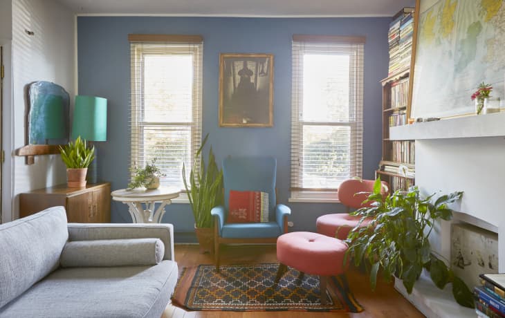 Cozy living room with blue walls, vintage armchair, pink ottomans, plants, bookshelves, and a framed portrait.