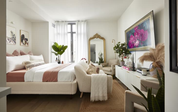 Studio apartment living and bedroom space with white walls, wood or laminate floor, neutral sofa, armchairs, bed with blush and white bedding, ornate gold mirror, large flower painting over white credenza, window with white sheer curtains