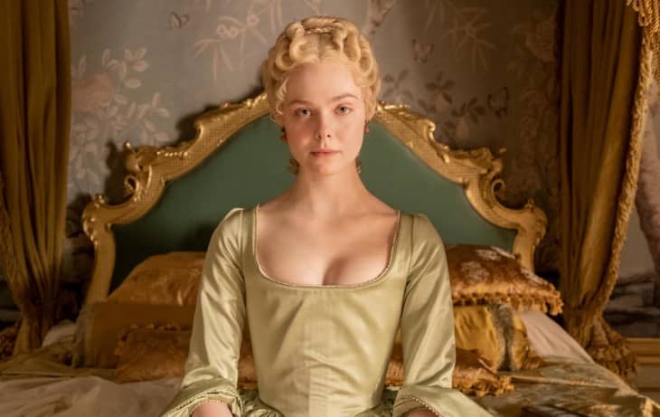 Woman in an ornate room wearing a green historical dress, sitting on a bed with a gold headboard and floral wallpaper.