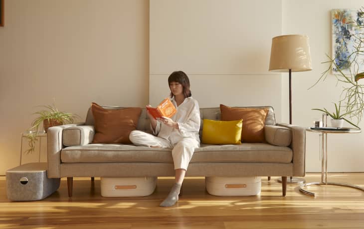 Woman reading on a beige sofa with orange and yellow cushions, surrounded by plants and a floor lamp.