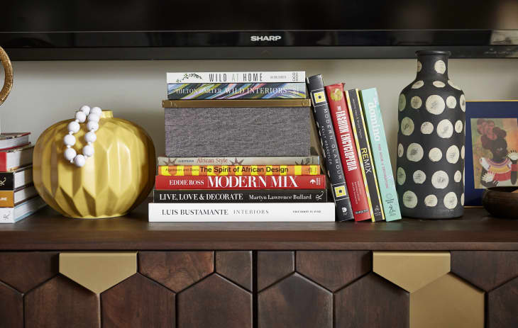 Books stacked on a wooden shelf with a yellow vase, black patterned vase, and framed art.