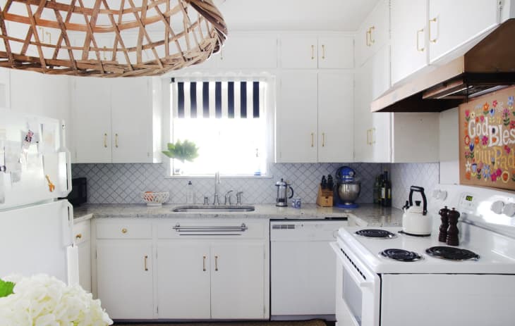 White kitchen with marble countertops, striped window shade, wicker light fixture, and appliances including a mixer and kettle.
