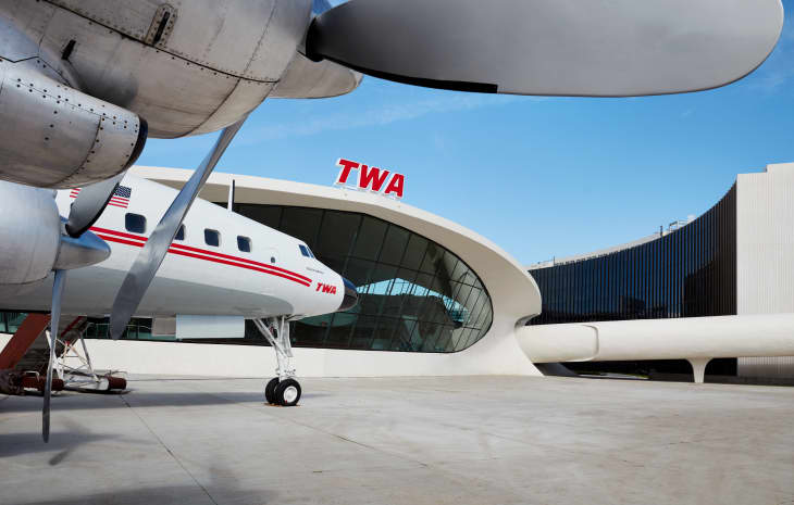 Vintage TWA airplane parked outside the TWA terminal with curved glass facade and red TWA sign.
