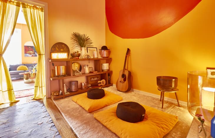 Meditation room with yellow cushions, a guitar, and a shelf with decor items, leading to a living room with yellow curtains.