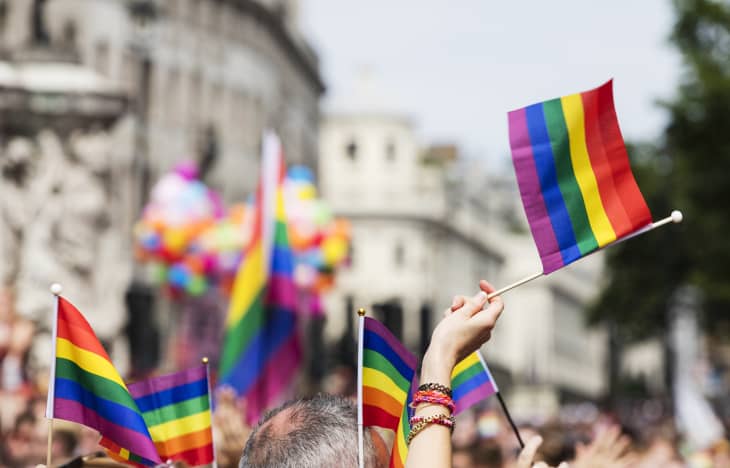 People holding rainbow flags at a pride parade with colorful balloons in the background.