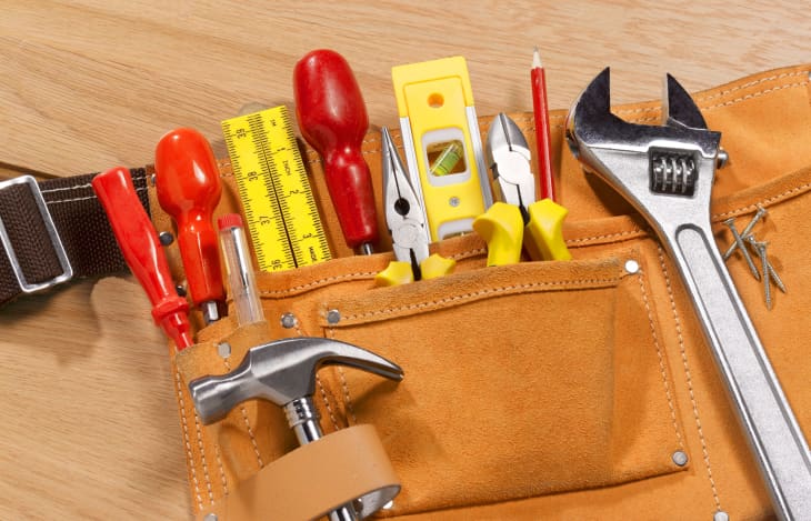 suede toolbelt on wood surface. full of yellow and red tools: wrench, hammer, pliers, level, ruler, screwdrivers