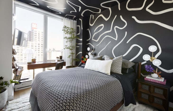 Bedroom with black walls with white bold design, bed has black and white bedding. There are large windows with a wood desk in front