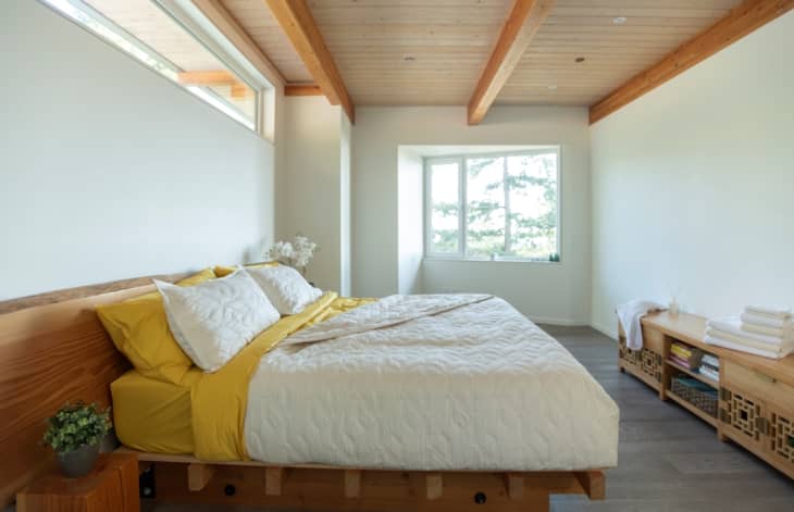 Minimalist bedroom with wooden beams, a bed with yellow sheets, white quilt, and a wooden bench with folded towels and books.