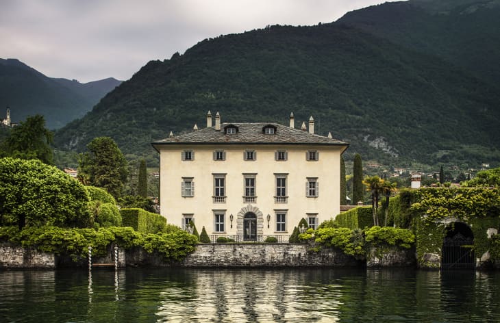 Elegant villa with arched windows and lush gardens by a lake, set against a backdrop of green mountains.