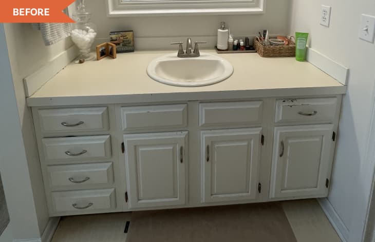 Bathroom vanity with white cabinets, single sink, and countertop items including a glass jar, framed photos, and toiletries.