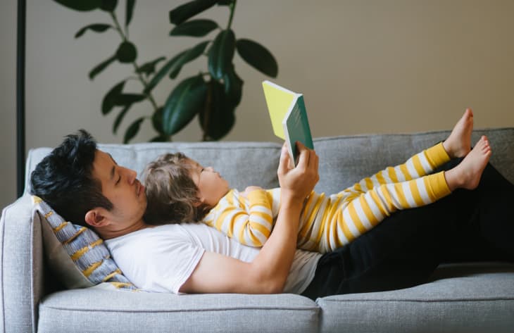 Man and child in yellow striped pajamas reading on a gray sofa with a large plant in the background.