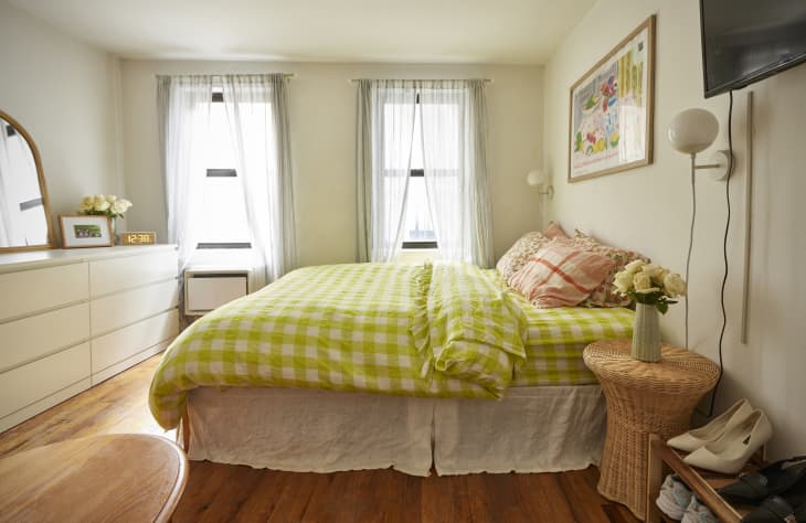 Bedroom with bed with chartreuse and white gingham duvet with floral and plaid pillowcases, straw round bedside table with case of yellow flowers, wood shoe rack with wall mount TV over it, windows with sheer white curtains, wood floor, white dresser with arched mirror across from bed