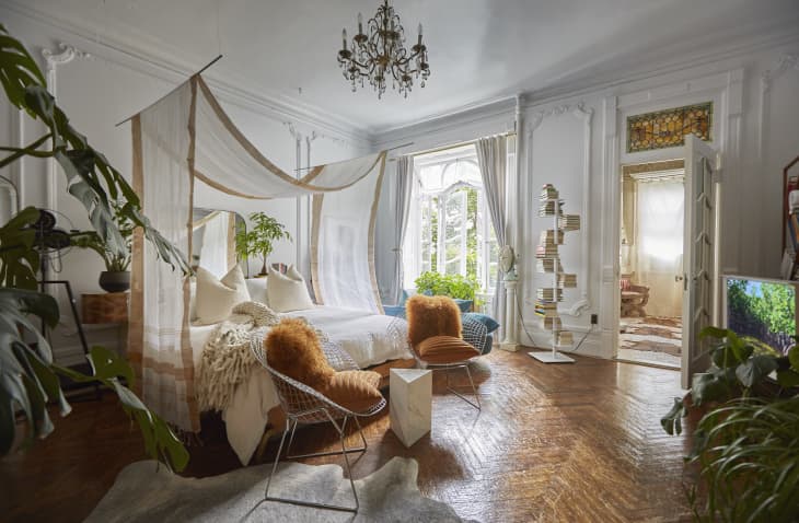 Elegant bedroom with canopy bed, chandelier, plants, and unique book tower near a large window.