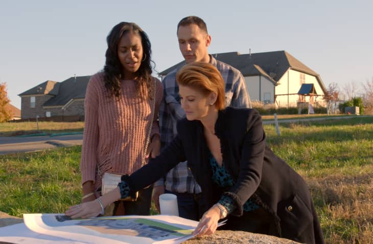 Three people reviewing architectural plans on a grassy plot with houses in the background.